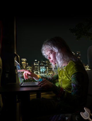 Eine Person sitzt auf dem Rücksitz eines Vans, durch die geöffnete Beifahrertür ist die Skyline einer Stadt bei Nacht zu sehen, ein MacBook Pro mit PROLED XDR Display auf einem Schreibtisch, die Person wird vom Bildschirm beleuchtet, während sie das Touch Feature nutzt, um ein Video in DaVinci Resolve Studio zu bearbeiten